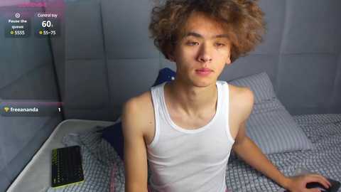 Media: Video of a slender, light-skinned, curly-haired young man with a neutral expression, wearing a white tank top, sitting on a bed with a checkered gray blanket. A black smartphone and blue pillow are visible.