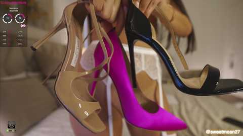 Media: Video of a woman holding two high-heeled shoes: beige with straps and a bright pink one with a pointed toe. Background shows a beige couch and a blurred, modern interior.