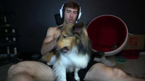 Media: Video of a shirtless man with headphones, sitting on the floor with a fluffy dog, in a dimly lit room with a red circular chair and dark walls.