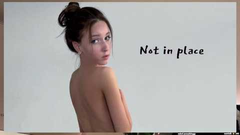 Media: Video of a slender, light-skinned young woman with dark hair in a bun, topless, looking over her shoulder. \"Not in place\" text is printed in black on a white background.