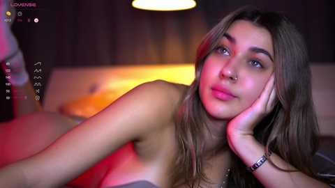 Media: A video of a young woman with fair skin and long brown hair, lying nude on a bed, wearing a bracelet. She has a thoughtful expression, with a hand under her cheek. The background is softly lit with warm hues.