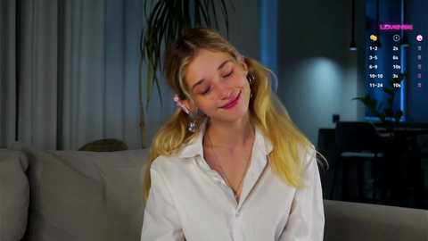 Media: Video of a smiling young woman with blonde pigtails, wearing white shirt, seated on a beige couch, in a dimly lit modern living room.