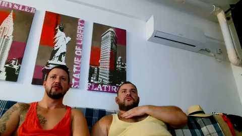 Media: Video of two men with tattoos, one in a red tank top, the other in a yellow tank top, sitting on a blue couch, in a room with a white wall, two framed art pieces, and an air conditioner.