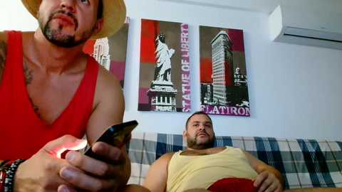 Media: Video of two men: a bearded, muscular man in a red tank top holding a phone, and a bald, heavier man in a yellow tank top, both on a plaid couch.