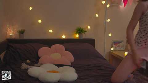 Media: Video of a cozy bedroom with a woman in a polka-dot nightgown standing near a bed adorned with large, plush flower pillows and fairy lights.