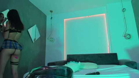 Media: A video shows a woman in a black bra and plaid skirt standing near a bed with neon lights, in a dimly lit bedroom.