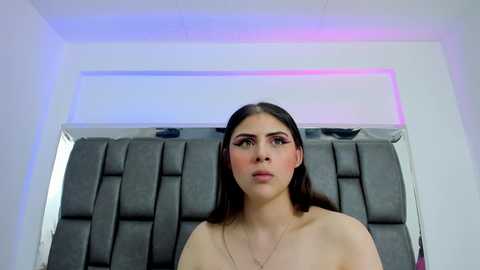 Media: Video of a young, light-skinned Latina woman with long, dark hair, wearing bold, pink eyeshadow and nude lipstick. She stands in front of a modern, grey upholstered headboard with blue and pink LED lighting.