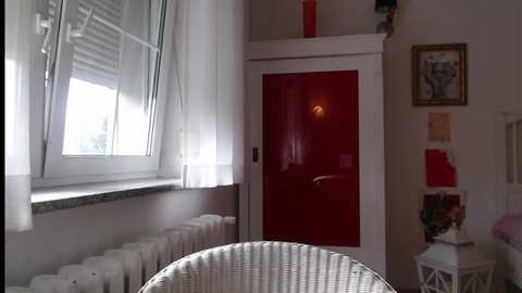 Media: A video of a modern, minimalist bedroom with white walls, a red door, and a white-framed window. A white wicker chair sits in the foreground.