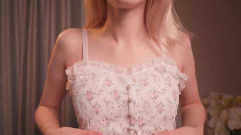 Media: Video of a fair-skinned woman with blonde hair, wearing a pastel floral dress with thin straps, standing in a softly lit room with muted curtains and a floral pillow in the background.