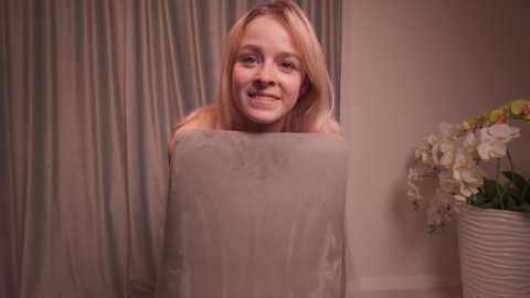 Media: Video of a smiling young woman with long blonde hair, wearing a beige sheet, standing in front of a white wall and floral arrangement.