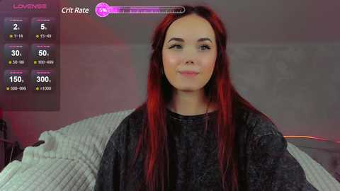 Media: Video of a fair-skinned woman with long, vibrant red hair, wearing a dark gray sweater, sitting on a bed with white sheets. In the background, a virtual game interface with player stats is visible.