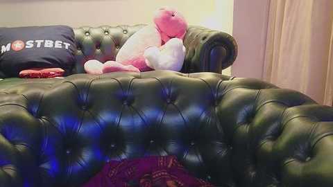 Media: Video of a plush green velvet sofa with tufted buttons, adorned with stuffed pink and white teddy bears, and a red pillow, against a beige wall and sheer curtains.