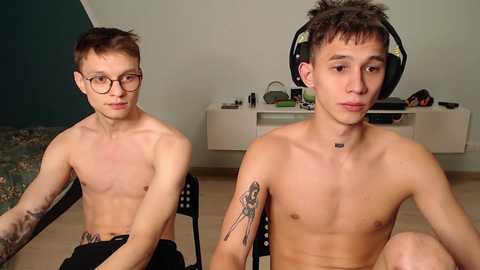 Media: Video of two young, shirtless men with pale skin and short dark hair. One wears glasses, tattoos, and sits on a chair, while the other has headphones, sits on a chair, and has a tattoo on his arm. Background features a plain room with a TV stand.