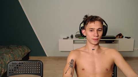 Media: Video of a shirtless, young, slim, Caucasian male with short brown hair, wearing large headphones, sitting indoors. He has a small tattoo on his upper left arm. Background includes a bed, a white shelf, and a green wall.