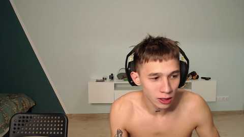 Media: Video of a shirtless young man with short brown hair, wearing large black headphones, sitting indoors. Background features a light green wall, white shelves, and a patterned couch.