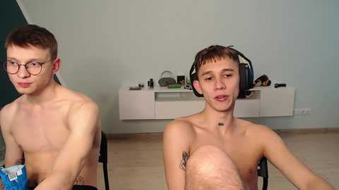 Media: Video of two shirtless young men, one with glasses and the other with headphones, seated on chairs in a minimalist room with white shelves and a blue wall.