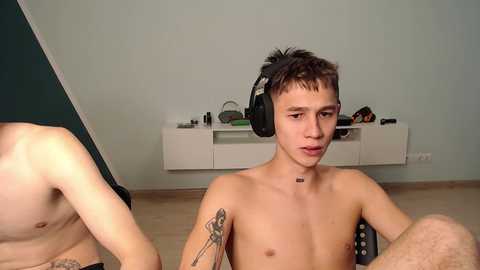 Media: Video of a shirtless young man with short dark hair, wearing headphones, seated indoors, with a tattoo on his left arm. Background shows a white shelf with items and light blue wall.