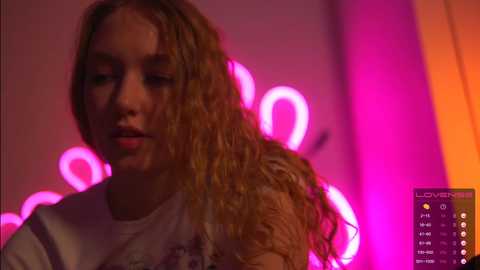 Media: Video of a young woman with wavy, blonde hair, wearing a white shirt, set against a neon pink and orange backdrop.