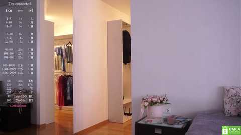 Media: Video of a modern, minimalist bedroom with a white wall, a partially open closet revealing clothes, and a pink flower arrangement on a black nightstand.