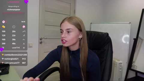 Media: Video of a young, light-skinned woman with straight, long brown hair, wearing a navy blue turtleneck, seated at a desk in an office. Background includes a whiteboard, door, and computer screen displaying social media notifications.