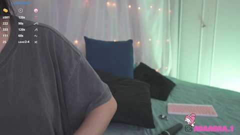 Media: Video of a person with short dark hair wearing a grey t-shirt, lying on a bed with blue pillows, surrounded by soft lighting and a pink keyboard on a desk.