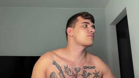 Media: Video of a shirtless, young white man with short dark hair, standing indoors. He has a muscular build and prominent chest tattoos including a detailed dragon and floral designs. The background is a simple, white-walled room with a dark TV on the wall.