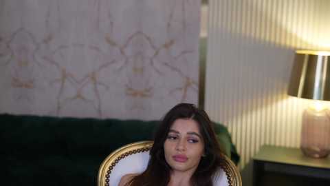 Media: Video of a young woman with long dark hair, sitting on a green velvet sofa with gold accents, in a warmly lit room with a beige floral-patterned wall and a lamp on a side table.