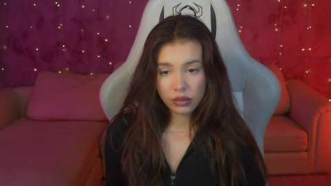 Media: Video of a young woman with long brown hair, wearing a black top, sitting in a plush, white gaming chair against a purple and red backdrop with twinkling lights.