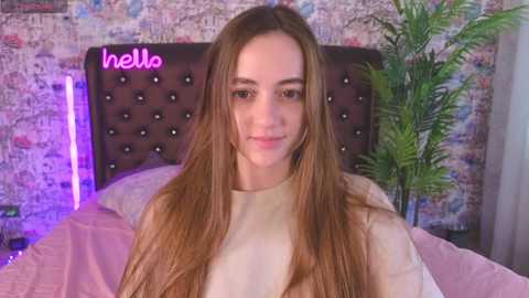 Media: Video of a young woman with long, straight, light brown hair, wearing a beige sweater, smiling in a bedroom with a tufted dark brown headboard, pink bedspread, and purple neon light.