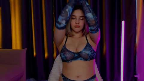 Media: Video of a young woman with light skin and shoulder-length brown hair, wearing blue lace lingerie, arms raised, in a dimly lit room with dark curtains.