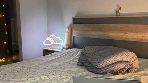 Media: A cozy bedroom with a wooden headboard, a bed with a gray patterned duvet, a bedside table, a neon cloud lamp, and a stuffed animal on the wall.