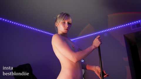 Media: Video of a topless, blonde woman with a slender physique, holding a microphone, in a dimly lit room with blue LED lights.