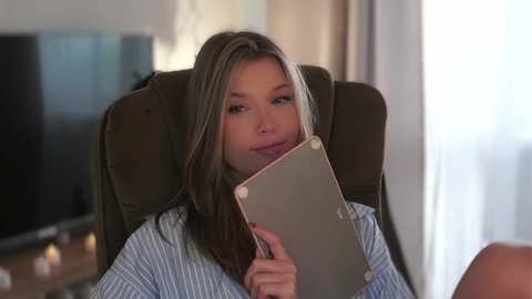 Media: Video of a blonde woman with light skin, wearing a blue striped shirt, holding a tablet, sitting on a brown armchair in a softly lit, modern living room.