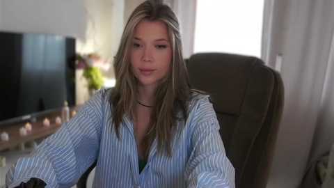 Media: Video of a young Caucasian woman with straight, light brown hair, wearing a blue and white striped button-down shirt, seated at a desk in a softly lit room with a TV and plants in the background.