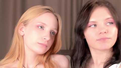 Media: Video of two young women with pale skin, one with long blonde hair, the other with long dark hair, both wearing pink eyeshadow, looking contemplative, background blurred.