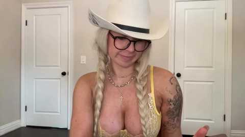 Media: Video of a middle-aged woman with long blonde hair, wearing a white cowboy hat, black-rimmed glasses, and a yellow lace bra, standing in front of white doors.