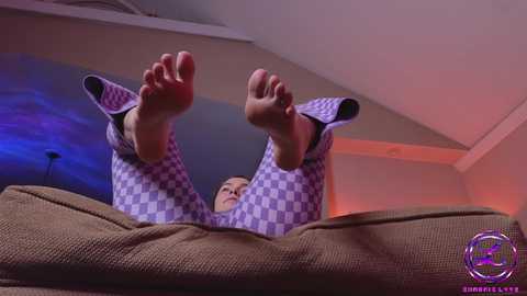 Media: Video of a woman lying on a brown couch, wearing purple checkered pajamas, with her feet raised towards the camera, against a dimly lit background with purple and blue lighting.