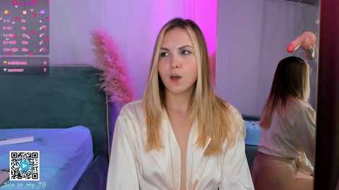 Media: Video of a blonde woman in a white silk robe, sitting on a bed in a dimly lit room, with a pink and blue light setting.