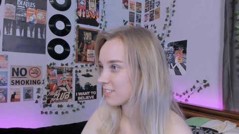 Media: Video of a young woman with long, platinum blonde hair, wearing a light-colored top, sitting in a room decorated with posters, including The Beatles and Led Zeppelin.