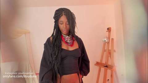 Media: A video shows a young woman with long, black braids, wearing a black, off-shoulder crop top and a red scarf, standing in a dimly lit room with a wooden easel and painting supplies in the background.