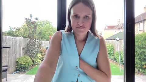 Media: Video of a fair-skinned woman with straight brown hair, wearing a light blue sleeveless blouse, standing in a bright, sunlit garden with greenery and a wooden fence in the background.