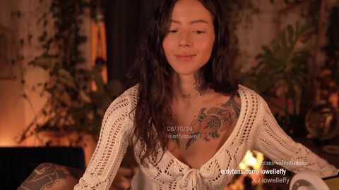 Media: Video of a smiling woman with long dark hair, wearing a knitted cream sweater, showcasing a colorful tattoo on her left shoulder, set against a warmly lit, bohemian interior with greenery and candles.