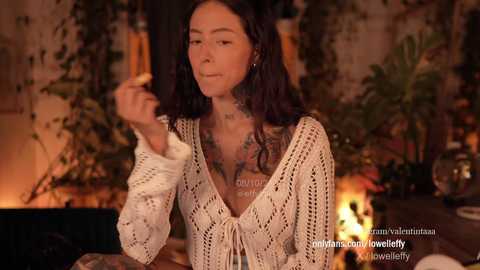 Media: Video of a tattooed woman with long dark hair, wearing a white crochet top, standing in a cozy room with warm lighting, surrounded by plants and candles.