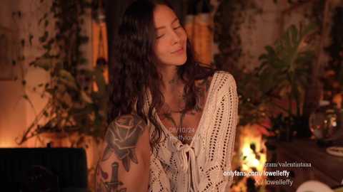 Media: Video of an Asian woman with long, wavy black hair, wearing a beige crochet top, adorned with tattoos. She stands in a warmly lit, bohemian room with greenery and potted plants in the background.