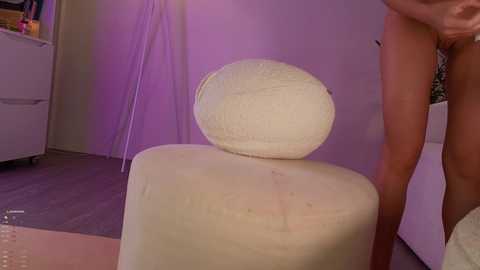 Media: A video of a naked person with fair skin, standing beside a white, spherical cake with a smooth texture, in a dimly lit room with purple lighting.