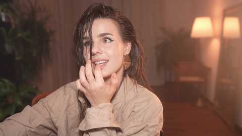 Media: Video of a smiling woman with dark hair, wearing a beige jacket, touching her face in a dimly lit room with a lamp and plant in the background.