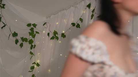 Media: Video of a woman with shoulder-length dark hair, wearing a white floral off-shoulder top, standing in front of a white curtain adorned with green ivy and fairy lights.