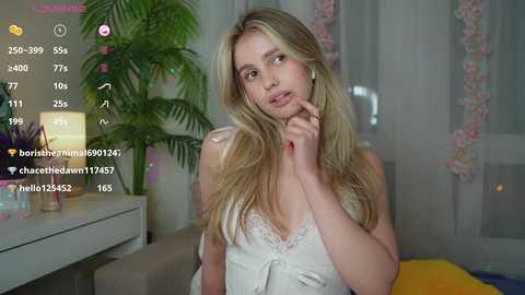 Media: Video of a blonde woman with fair skin, wearing a white lace camisole, sitting in a cozy room with a green plant, pink flowers, and a yellow pillow in the background.