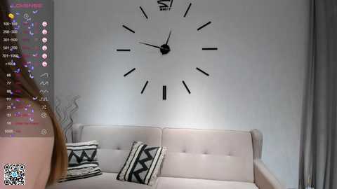 Media: A video of a minimalist living room with a beige sofa, black geometric pillows, and a large, minimalist clock on a white wall.