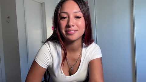 Media: Video of a young woman with medium skin tone, straight dark brown hair with red highlights, wearing a white top and a gold necklace, smiling slightly. She is indoors with white walls and a closed door in the background.